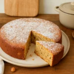 Photo d’un gâteau aux noisettes moelleux, fait maison, doré et saupoudré de sucre glace, présenté sur une table de cuisine avec des noisettes entières autour.