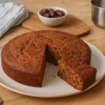 Photo d’un gâteau aux dattes moelleux et doré, fait maison, sur une assiette rustique, entouré d’ustensiles de cuisine – recette traditionnelle facile et parfumée, parfaite pour le Ramadan ou le goûter.