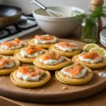 Blinis au saumon fumé garnis de crème fraîche et d’aneth, présentés sur une planche en bois, apéritif chic et facile pour les fêtes de Noël.