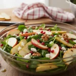 Salade de Noël festive aux épinards frais, pommes tranchées, noix croquantes, fromage émietté et graines de grenade, servie dans un saladier en verre sur une table en bois.